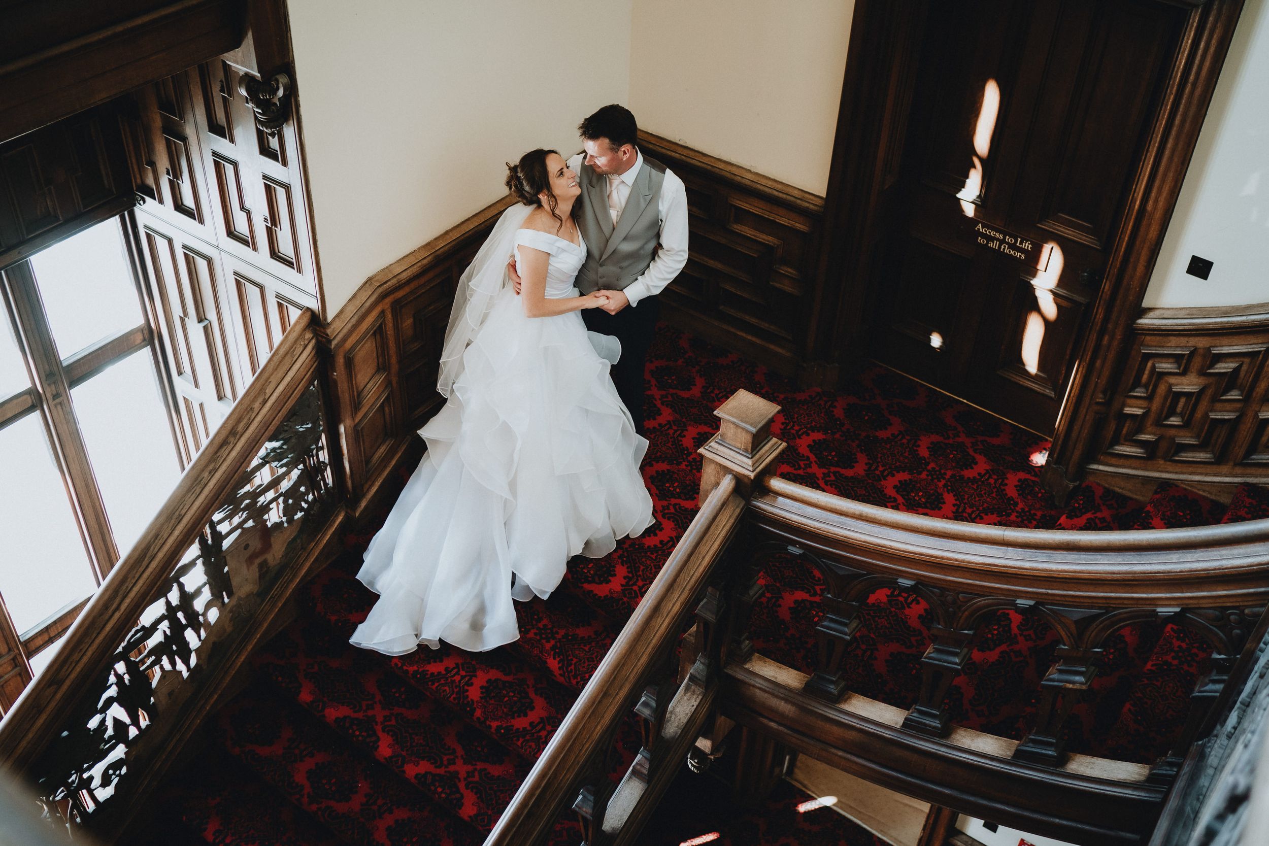 A couple in elegant wedding attire descend a grand staircase with intricate woodwork and red carpeting in a historic venue.