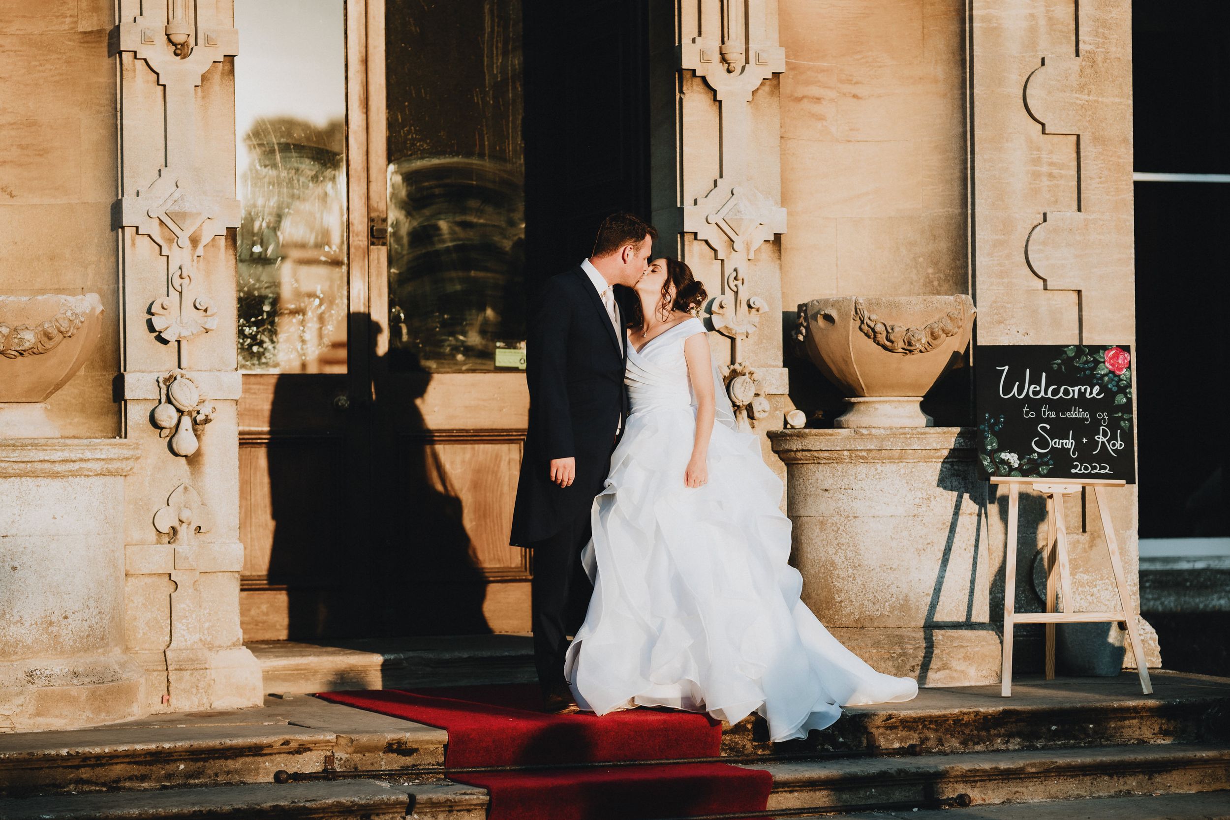 A couple in wedding attire stands on stone steps with a red carpet and ornate architectural details at a historic venue.
