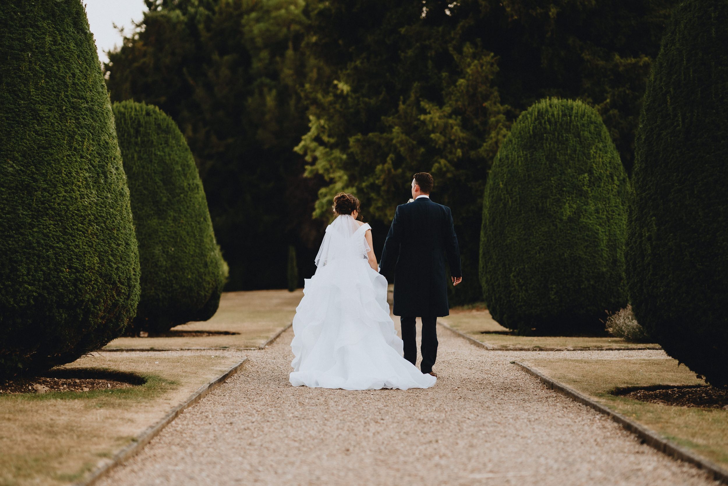 A couple in wedding attire walks down a formal garden path lined with perfectly manicured topiary shrubs.