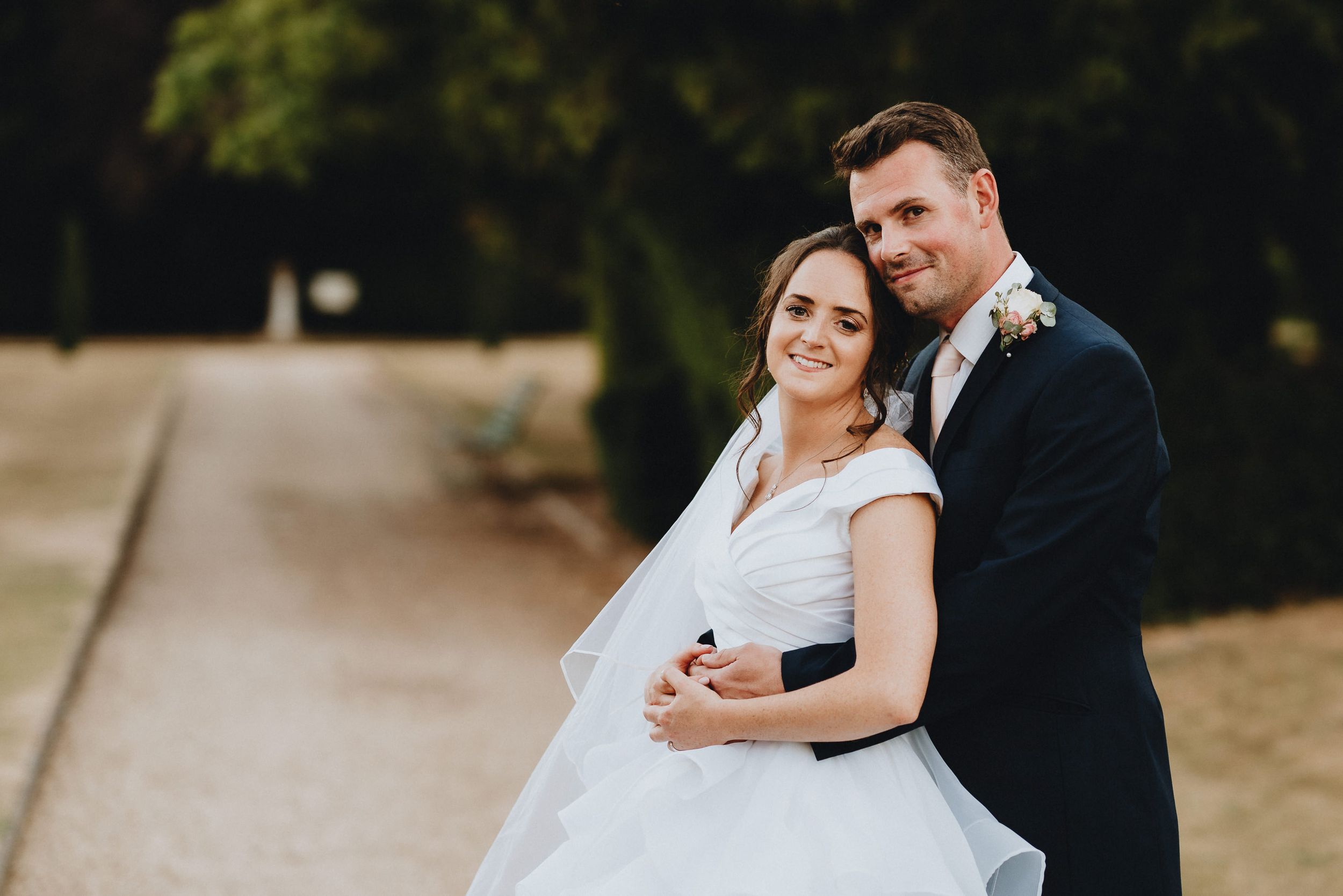 A bride and groom embrace on a scenic path during their wedding photo session in an outdoor setting.