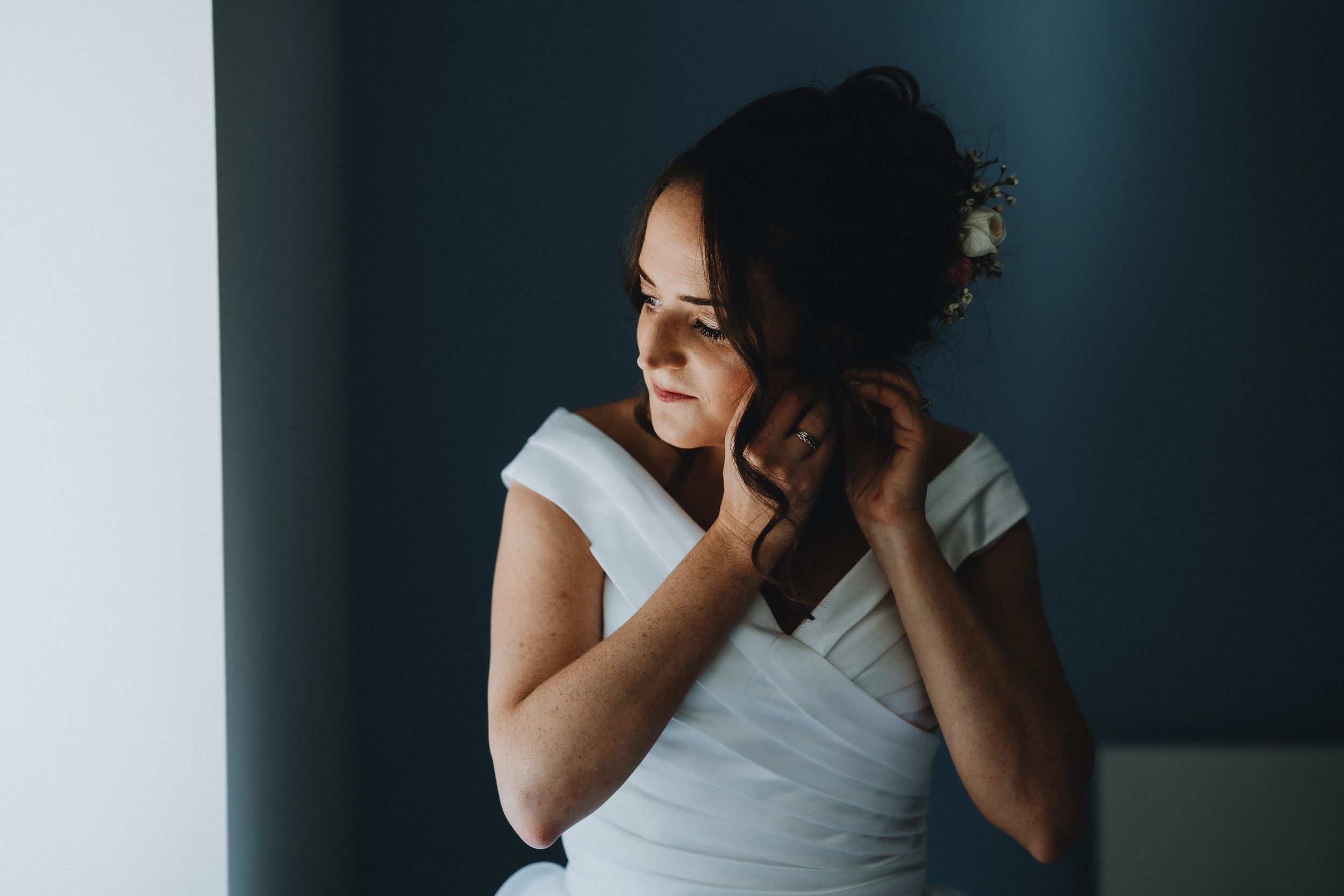 A figure in a white dress stands thoughtfully near a dark blue wall in moody lighting.