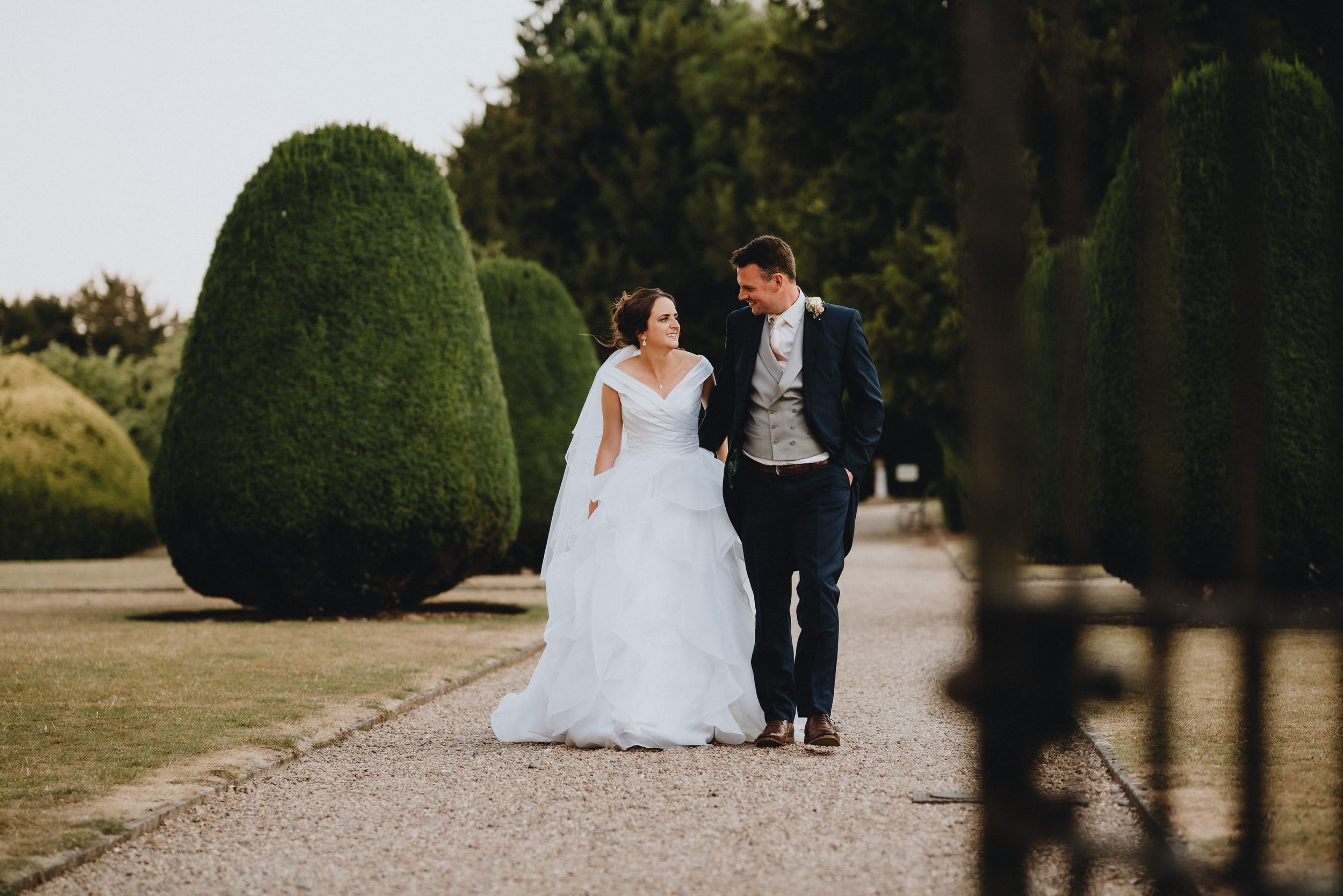 A couple in formal wedding attire walk together along a garden path at Stoke Rochford Hall lined with manicured topiary bushes at sunset.
