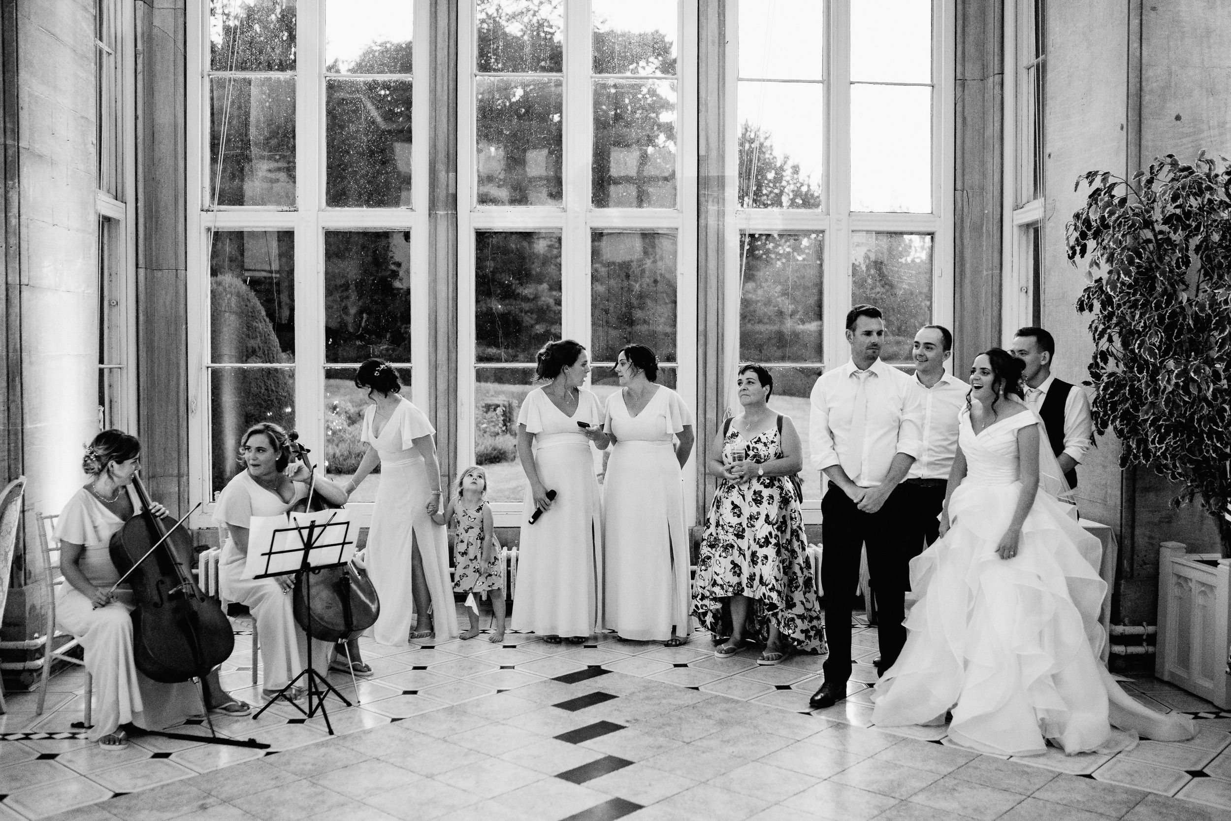 Black and white photograph of a formal celebration with musicians and people in formal attire inside an elegant room with windows.