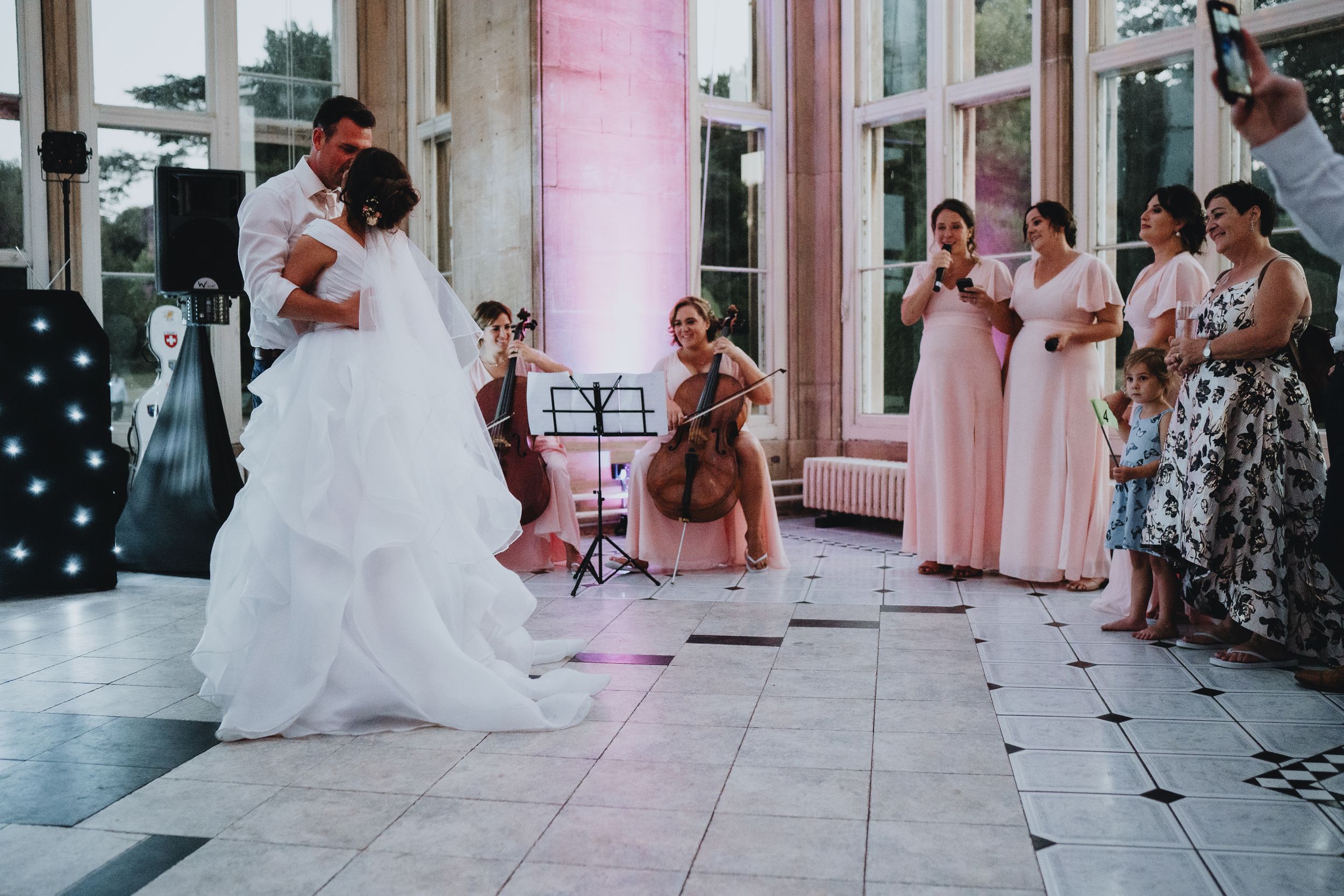 A couple dances at their wedding reception while guests in pink bridesmaid dresses and formal attire watch from the sidelines.
