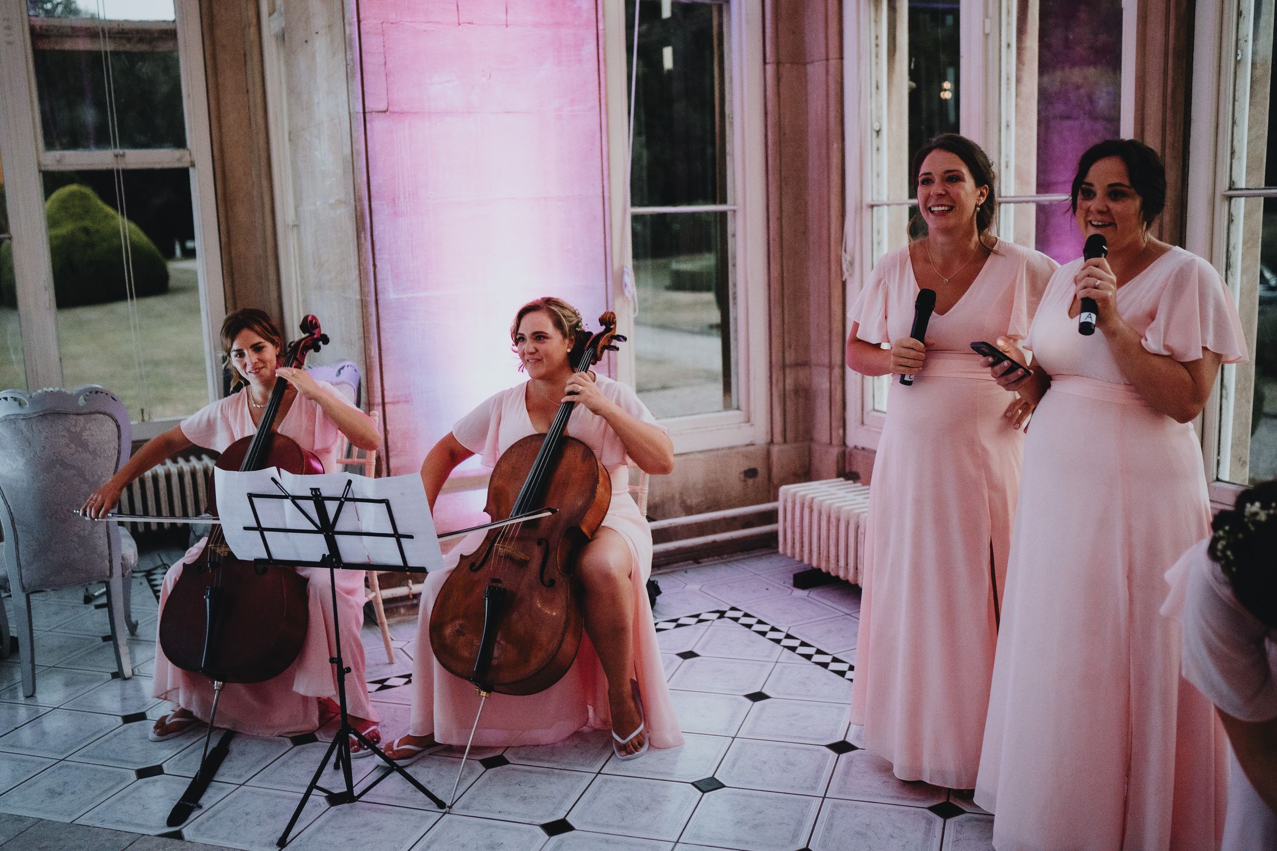 Musicians in pink dresses perform at an elegant venue with cellos and music stands.