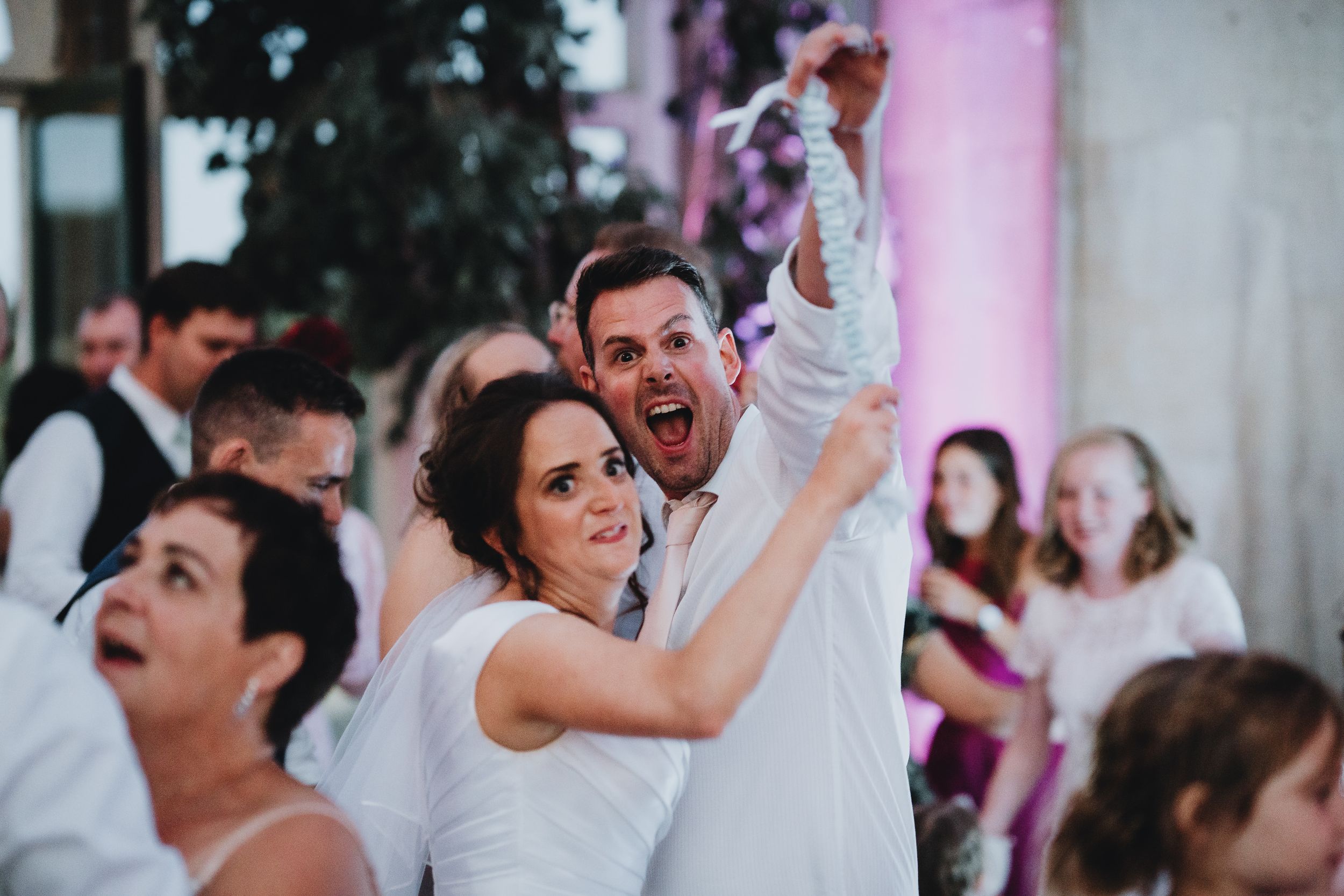 Wedding guests celebrate joyfully during a reception with arms raised in excitement at an indoor venue.