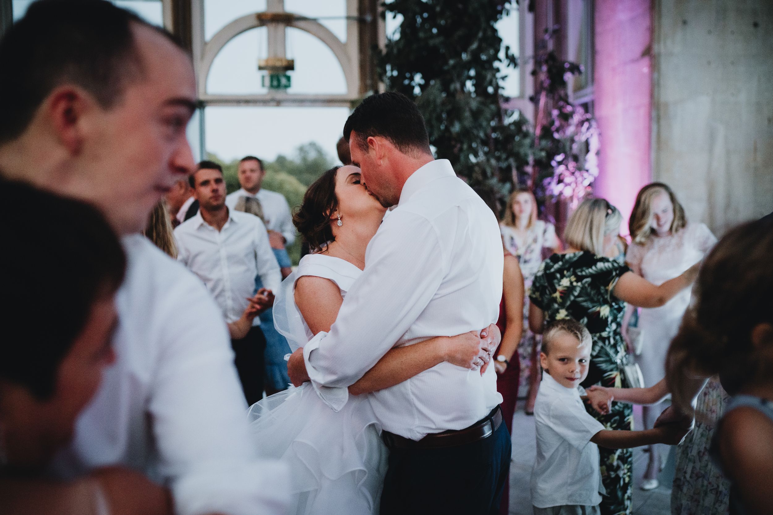 Intimate moment captured during wedding celebration with guests dancing in the background.