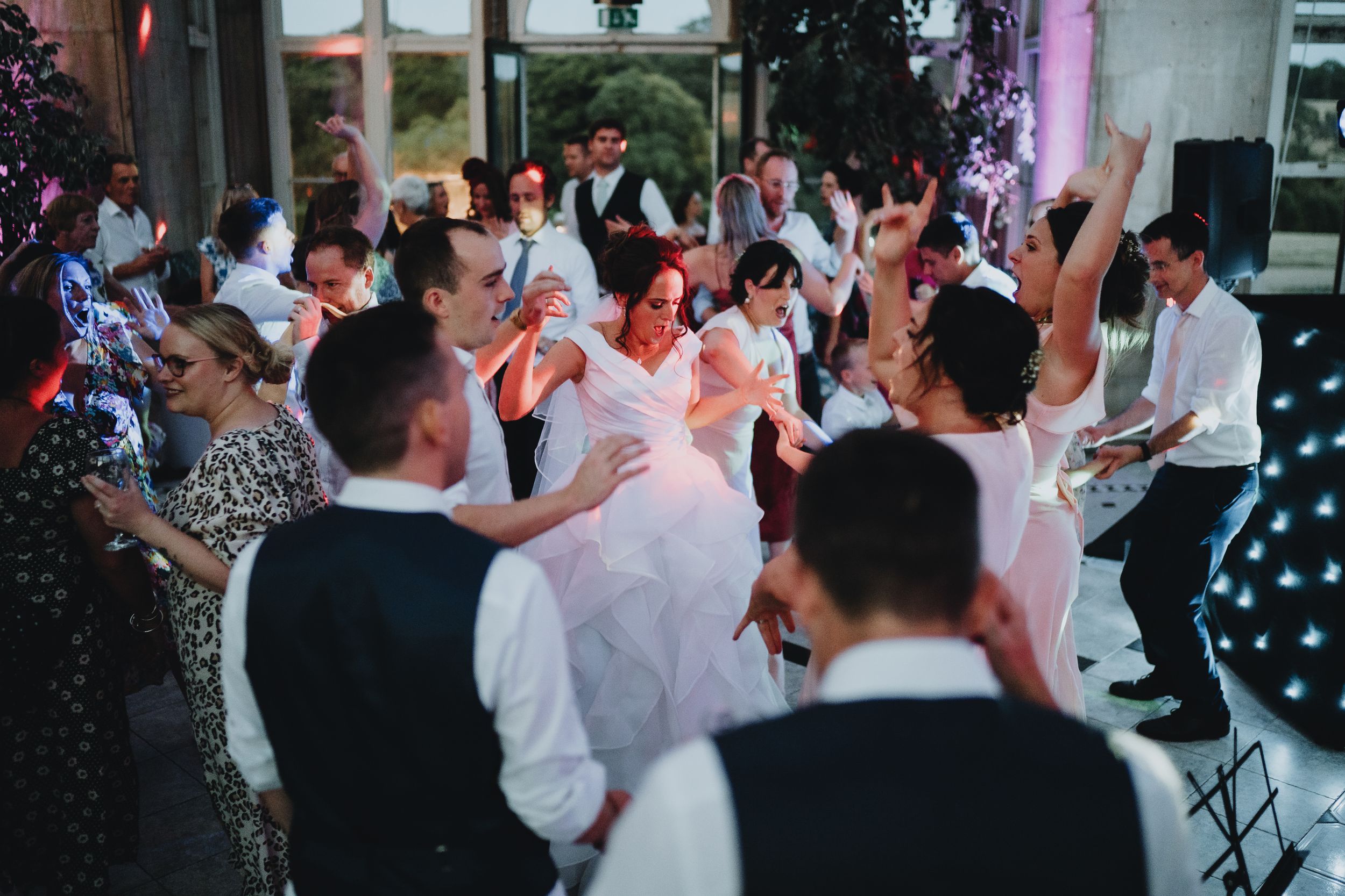 Party guests dance and celebrate on a crowded dance floor at an evening reception with colorful lighting.