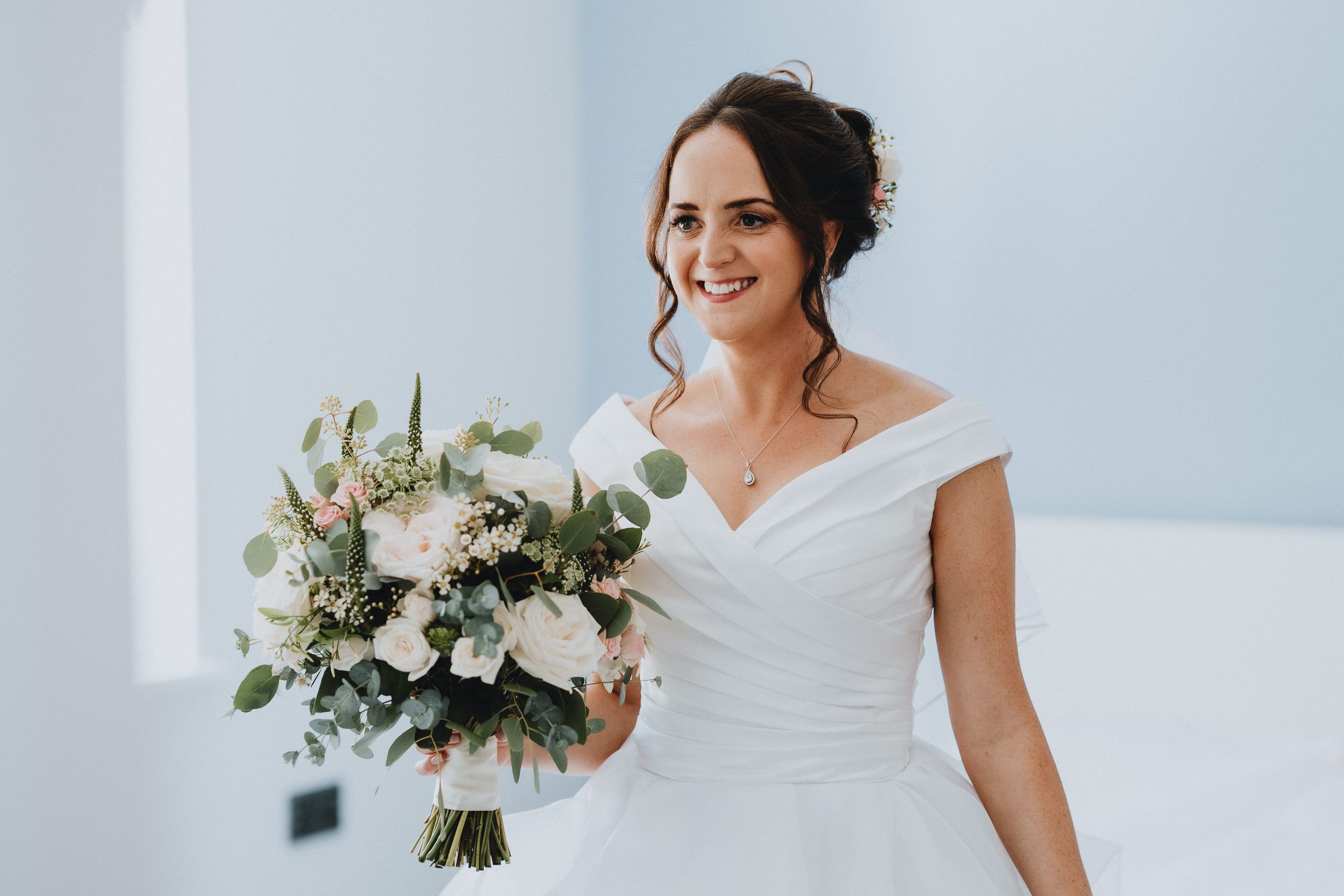 Beautiful wedding portrait with white off-shoulder gown and elegant bouquet of white flowers and greenery against blue wall.