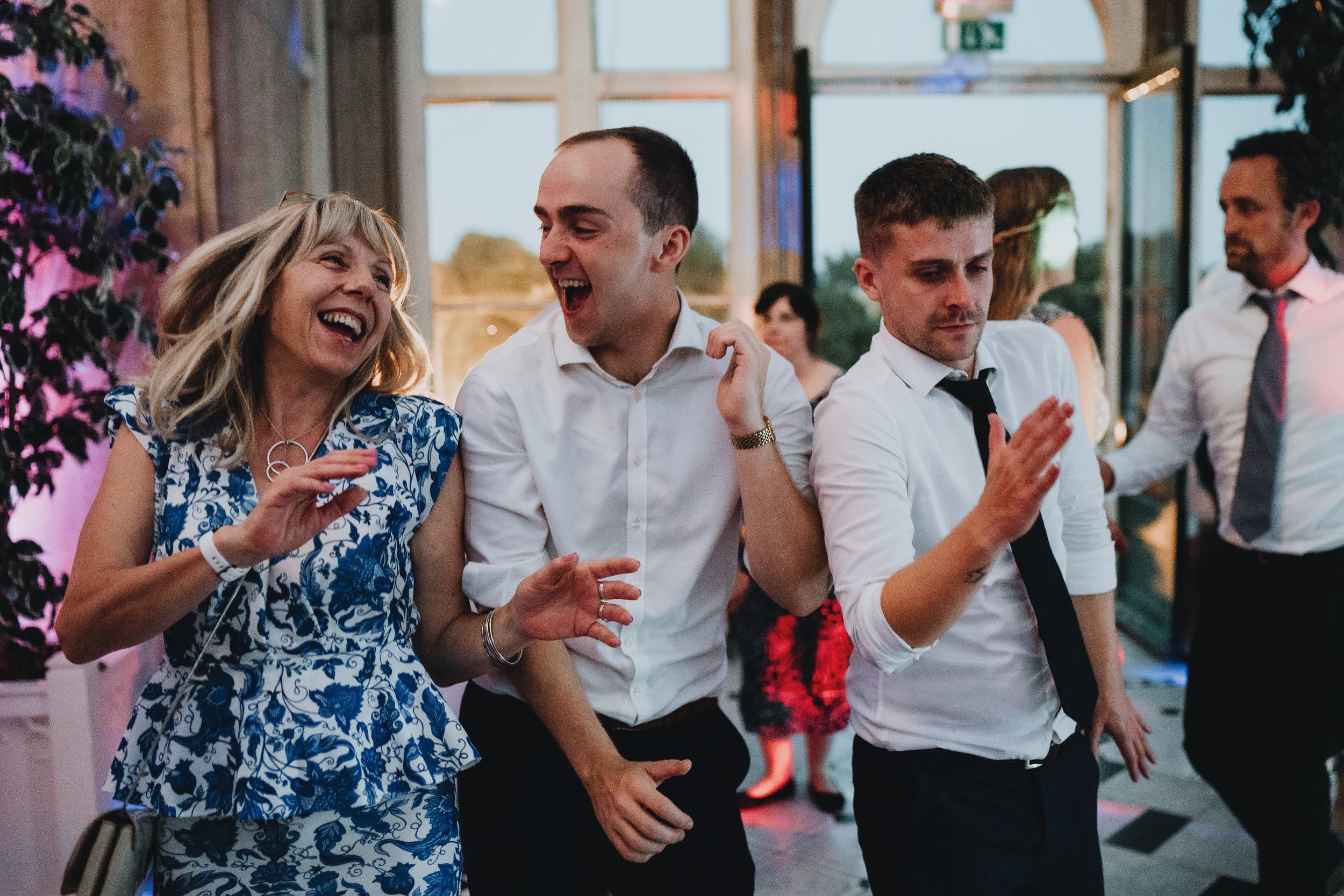 Wedding guests laugh and dance together at a reception with festive decorations visible in the background.