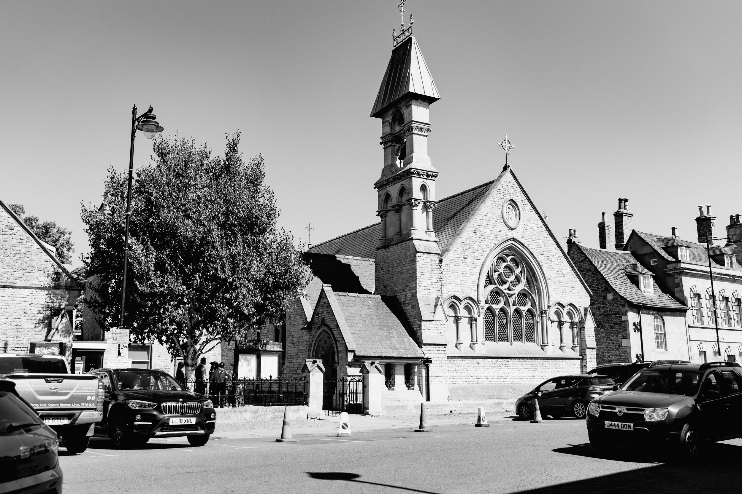 Black and white photograph of a historic church in Stamford with tall spire and arched windows surrounded by parked cars on street.