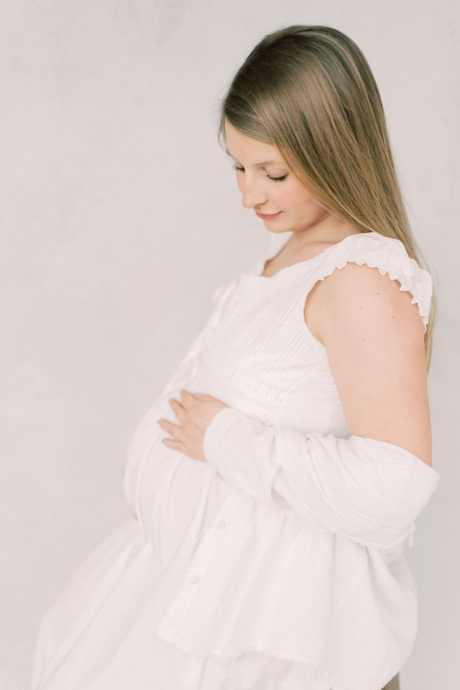 A pregnant woman in a white dress gently cradles her belly while looking down in a soft, ethereal maternity photo.