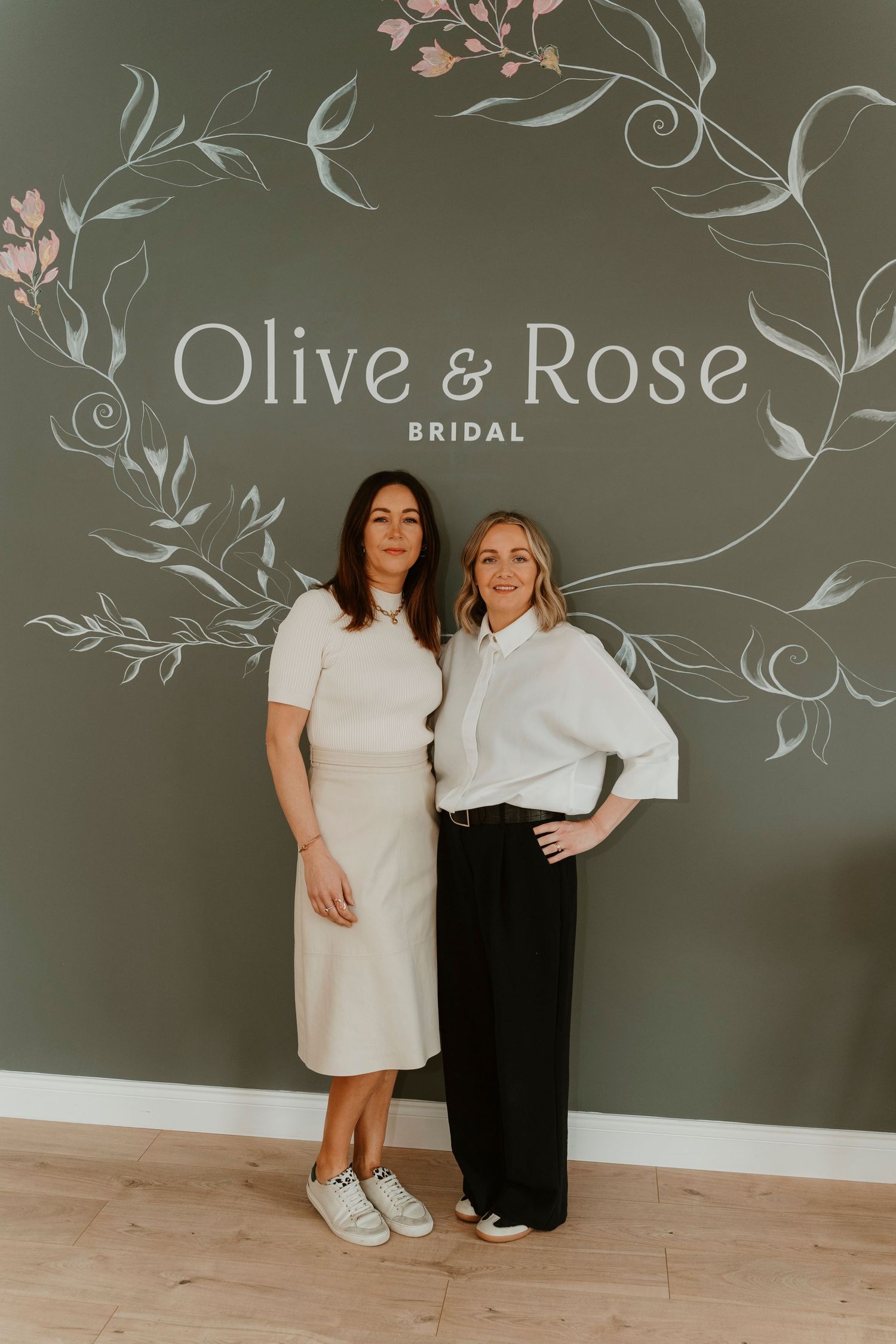 Two people stand in front of an elegant Olive & Rose Bridal logo wall wearing white tops and contrasting bottoms.