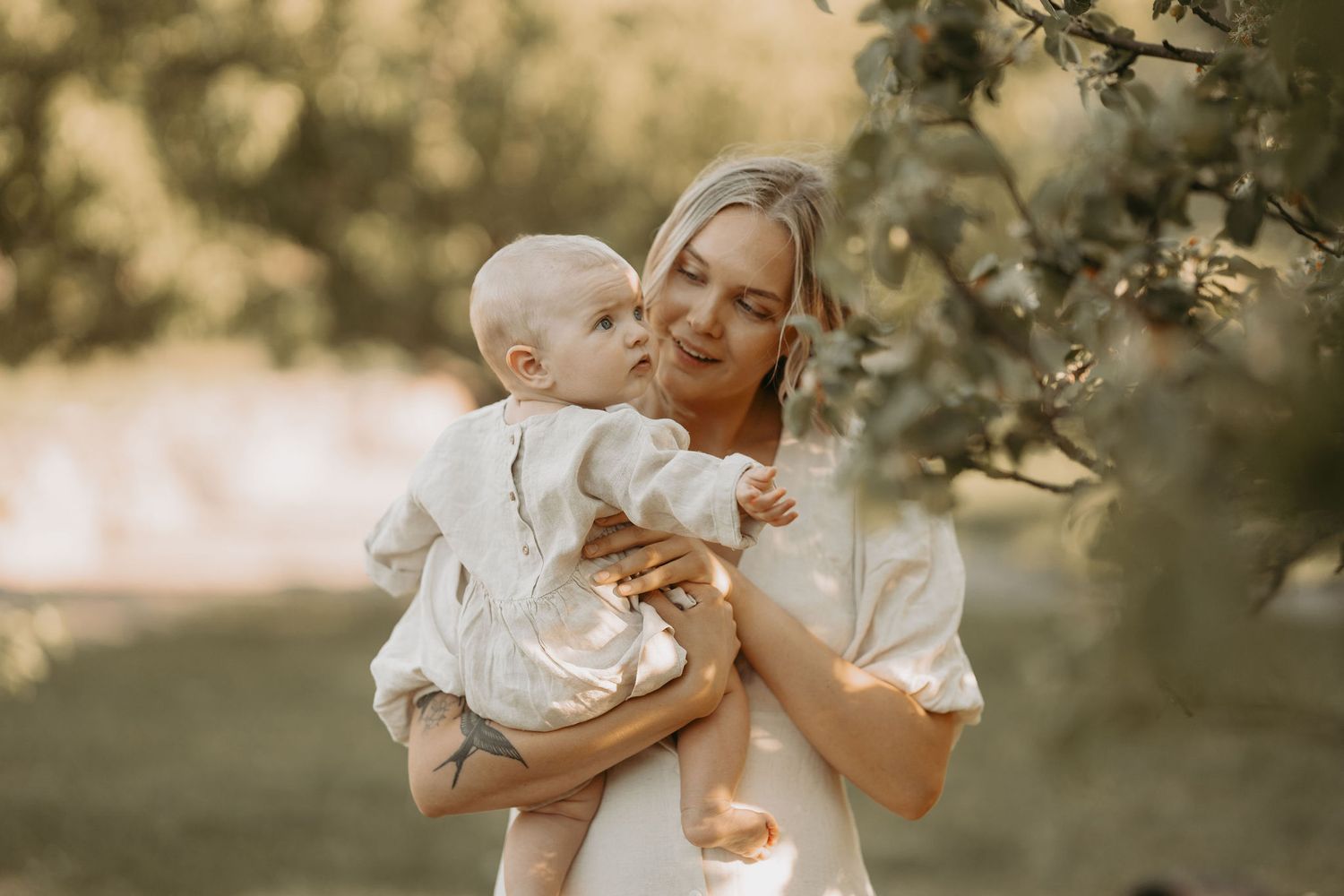 A tender moment captured in warm sunlight as parent and baby in white outfits share an intimate outdoor embrace.