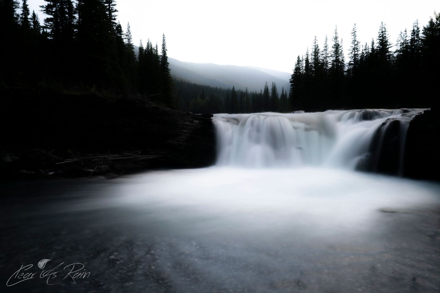 A long exposure waterfall cascades through evergreen forest in misty mountain landscape during twilight.