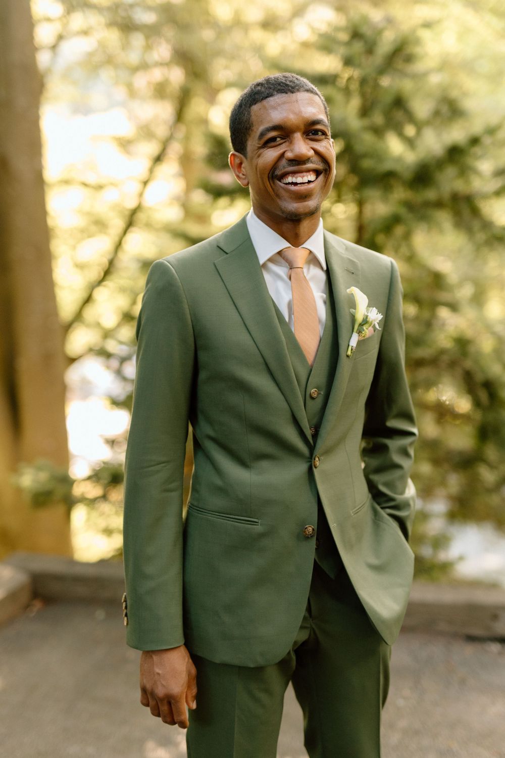 A groom wearing an olive green three-piece suit with peach tie smiles warmly outdoors in natural lighting.