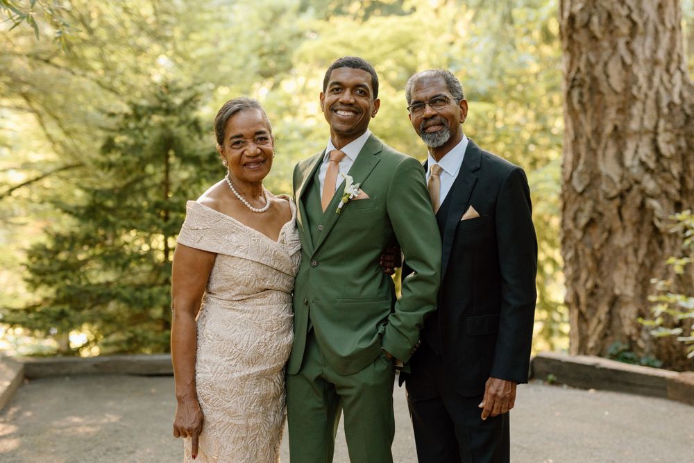 Three people in formal attire pose together outdoors, with one wearing a green suit and others in beige and black.