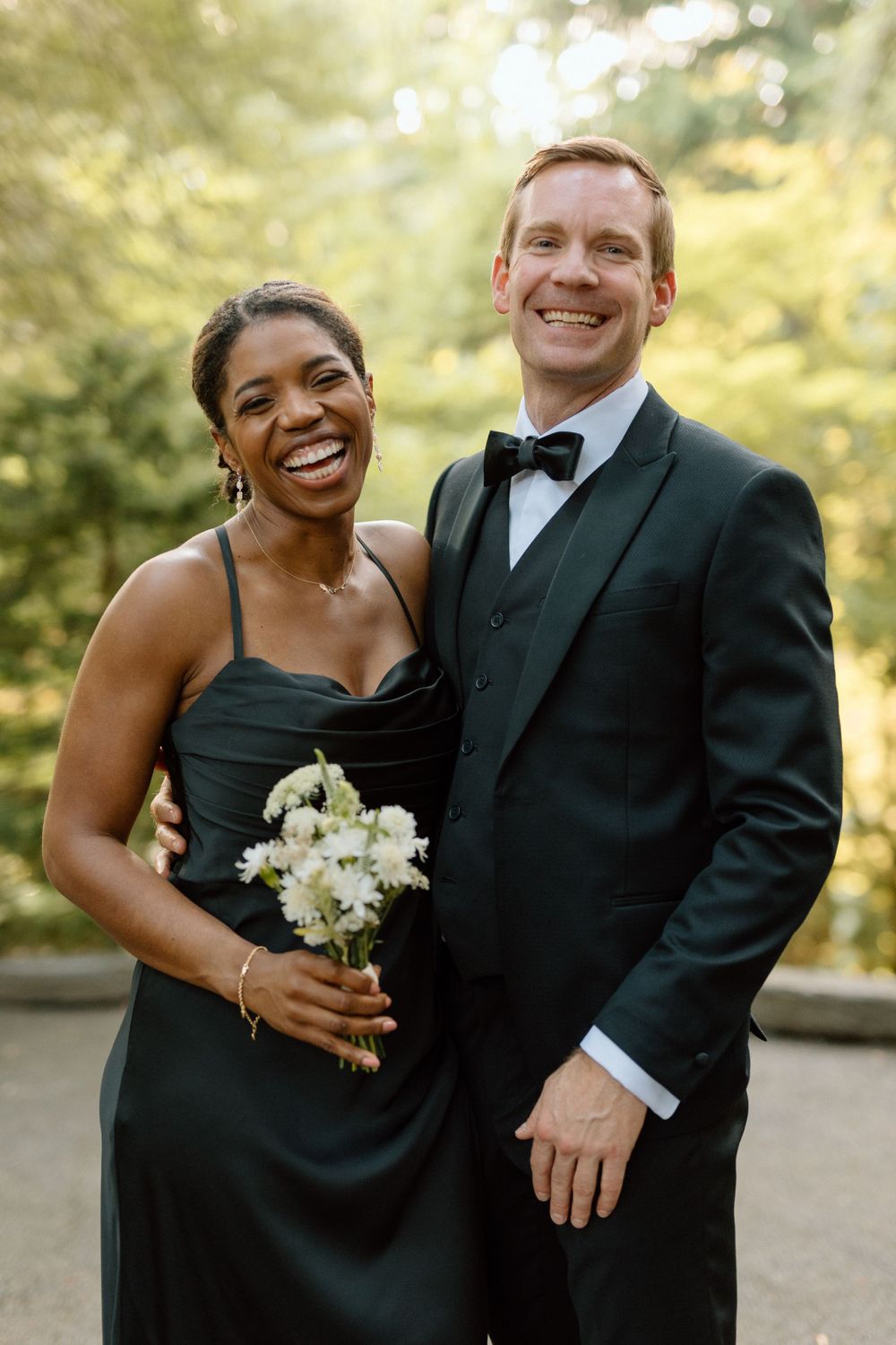A joyful couple in wedding attire smile together outdoors as they pose with a white floral bouquet.