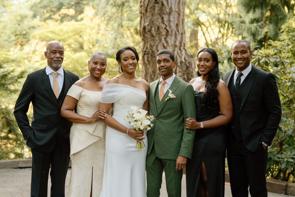 A wedding party poses together outdoors in formal attire, with the couple in white and green suits holding a bouquet.