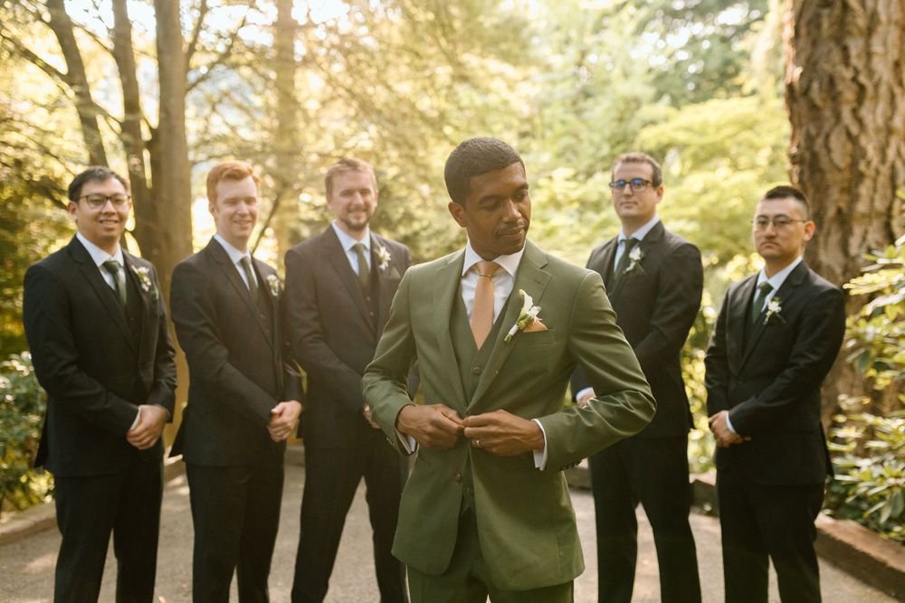 Wedding party with groomsmen in black suits and one in green standing outdoors in a sunlit forest setting.
