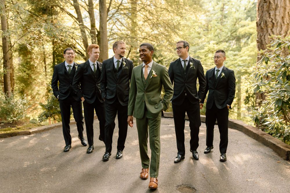 A group of groomsmen in black suits walk down a tree-lined path behind someone wearing a distinctive olive green suit.