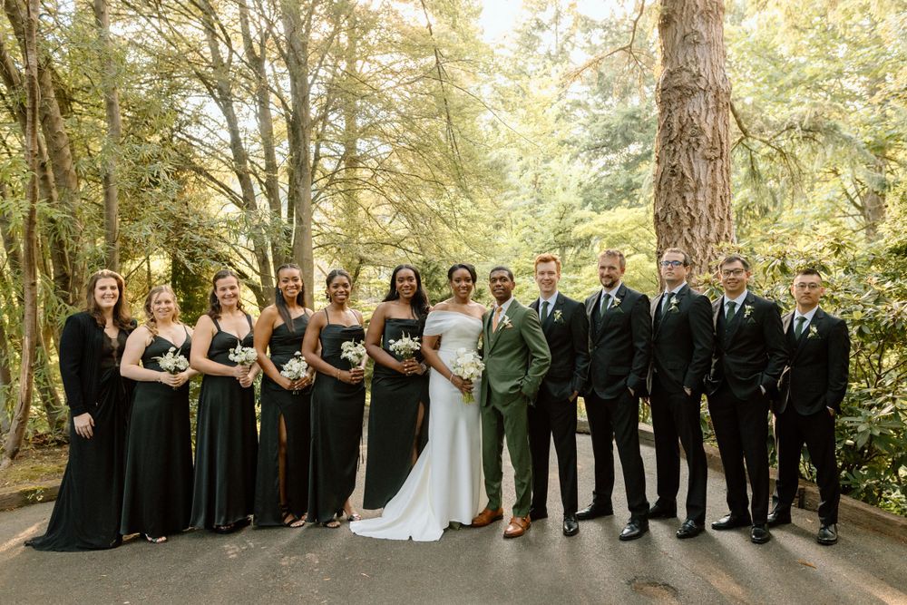 A wedding party wearing black and green formal attire poses together on a forested path beneath tall trees.