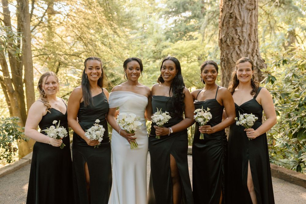 Wedding party wearing contrasting black and white gowns hold white flower bouquets on a wooded path.