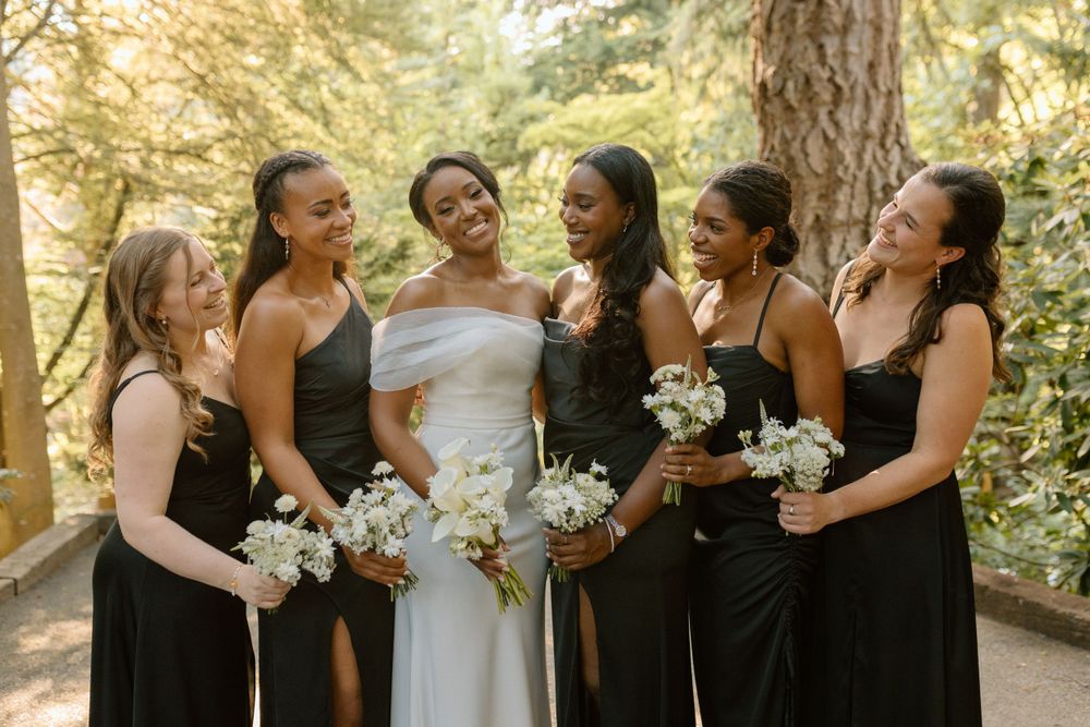 A wedding party wearing black bridesmaid dresses and white bridal gown hold bouquets while sharing a joyful moment outdoors.
