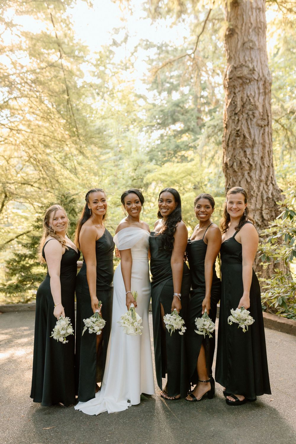 A bridal party in black bridesmaid dresses and a white wedding gown pose together on a tree-lined path.