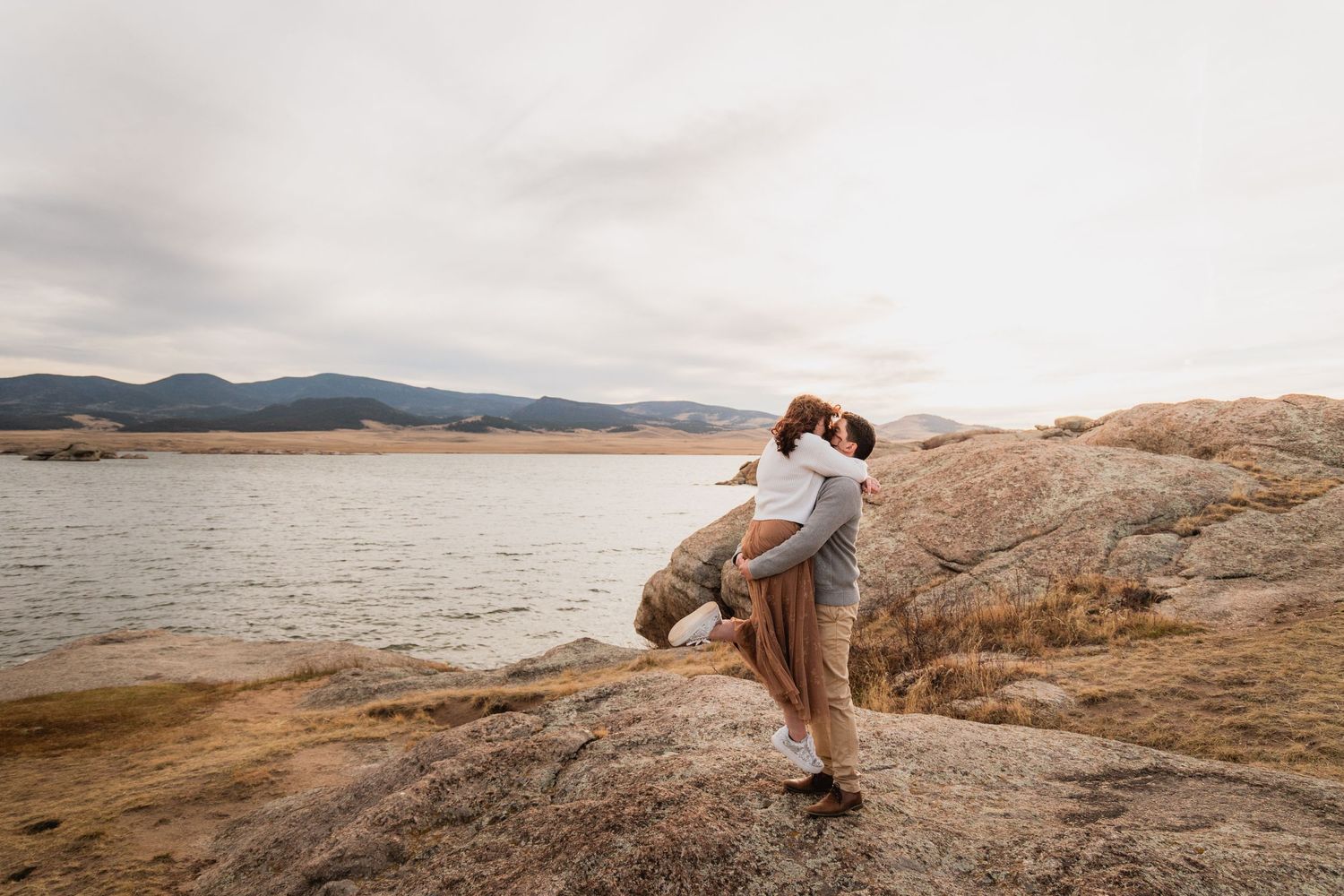 Couple embracing on rocky outcrop overlooking serene lake with mountains in the background at sunset.