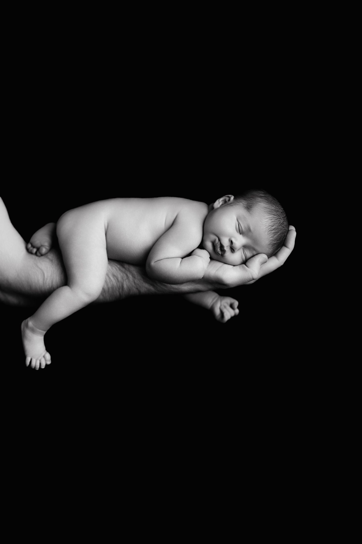 Black and white photograph of a sleeping newborn baby curled up peacefully against a dark background.