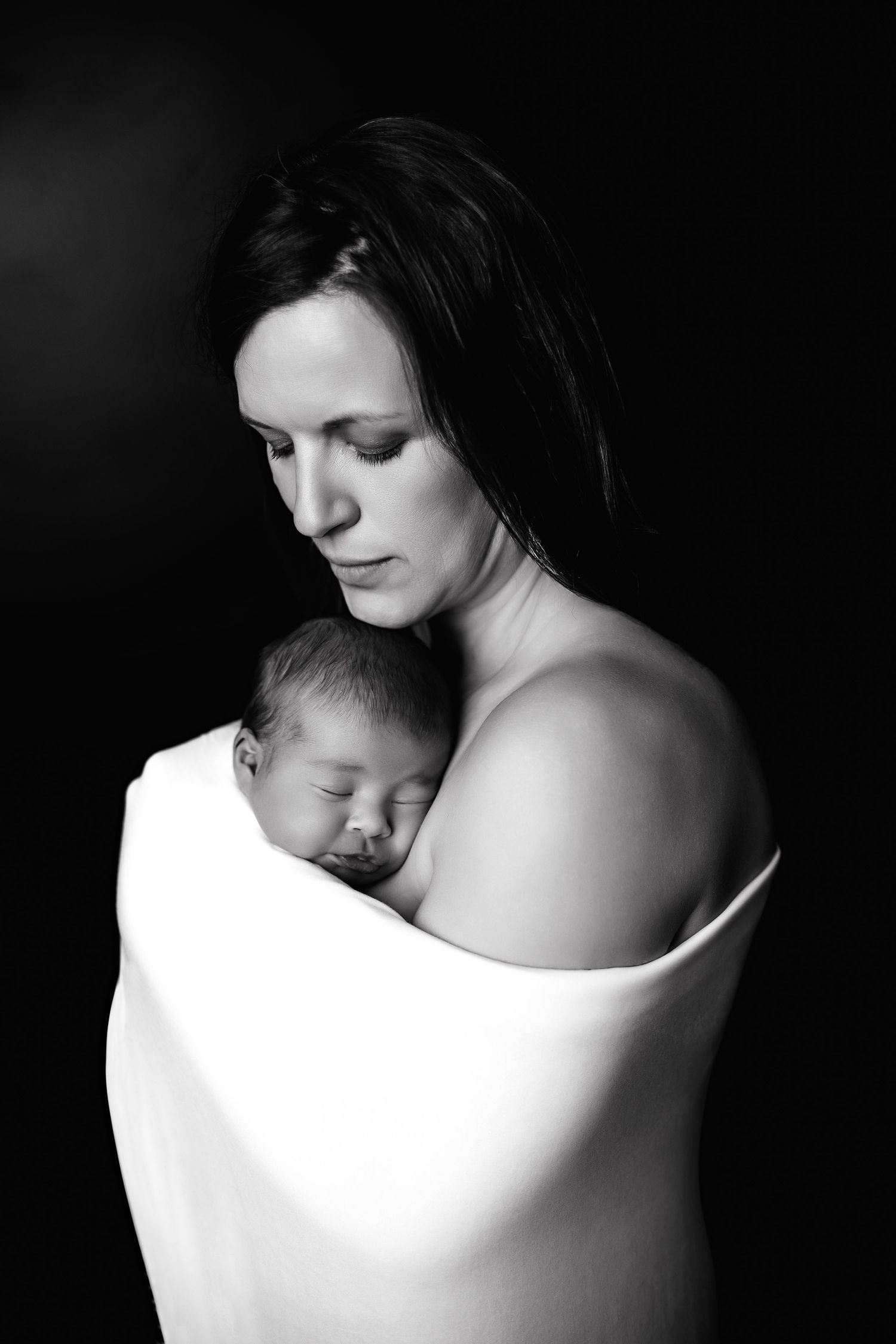 A tender black and white portrait of mother and sleeping newborn baby wrapped together in white fabric against dark background.
