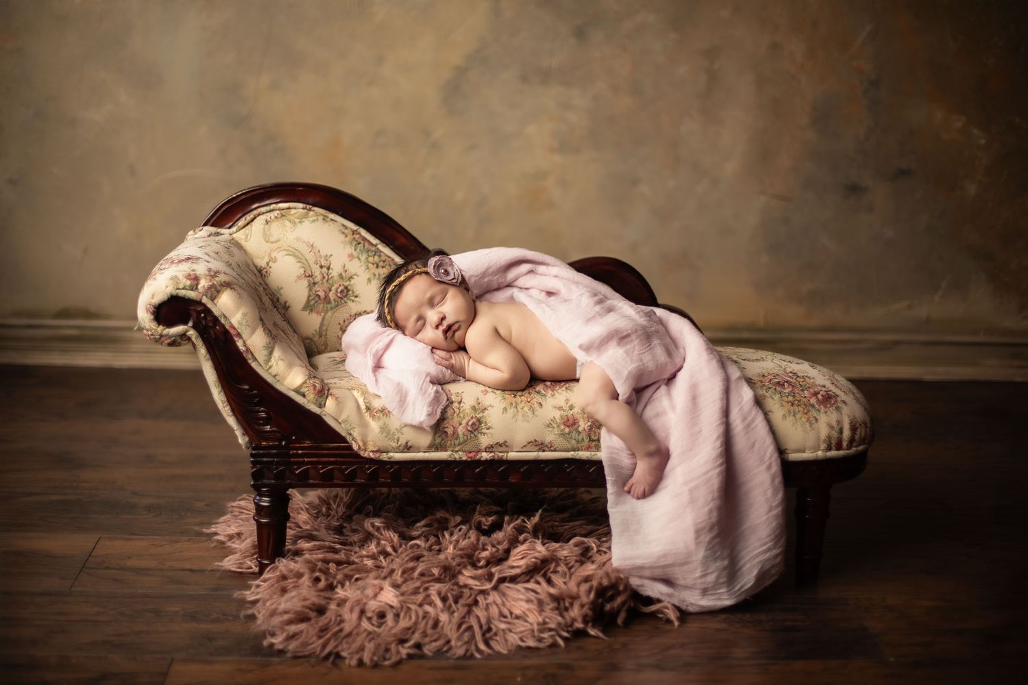A newborn baby sleeps peacefully on an antique chaise lounge draped with soft pink fabric and plush fur rug.