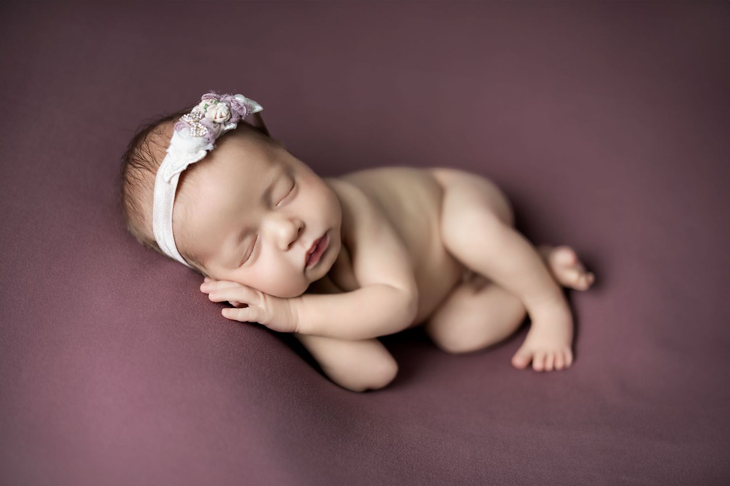 A peaceful sleeping newborn wearing a delicate white lace headband poses on a mauve background.