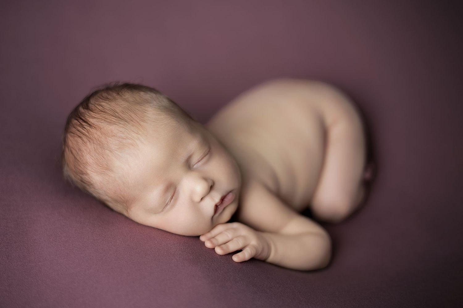 A sleeping newborn baby curled up peacefully on a purple backdrop in a soft, artistic pose.