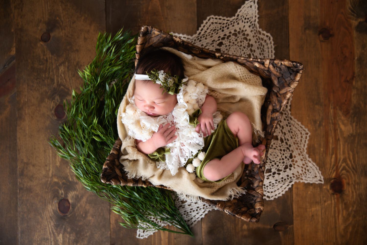 Overhead view of sleeping newborn on rustic wood floor with green wreath and lace decoration.