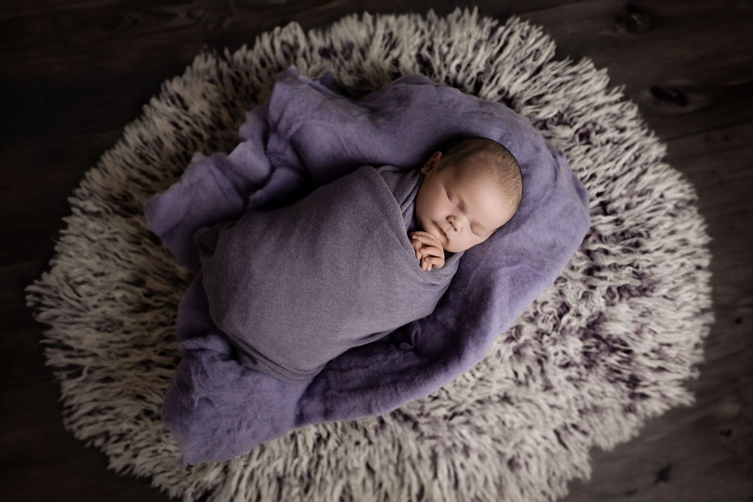 Newborn baby peacefully sleeping swaddled in purple fabric on a fluffy white circular rug.