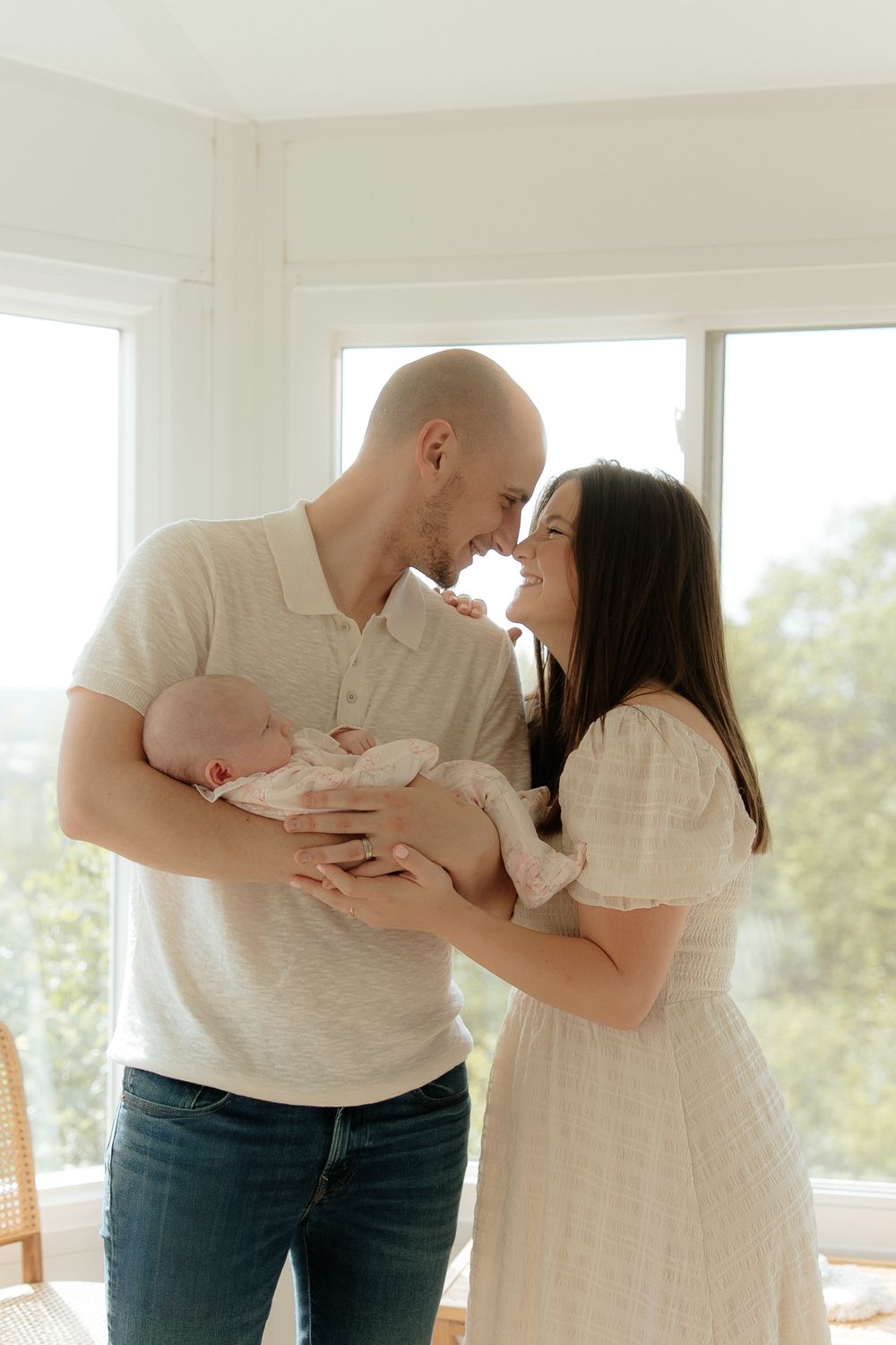 Couple in white clothing shares intimate moment while holding newborn baby beside bright windows in sunlit room.