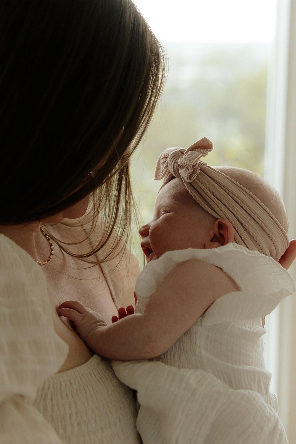 A tender moment captured of a parent holding a sleeping newborn in white clothing against a soft window light backdrop.