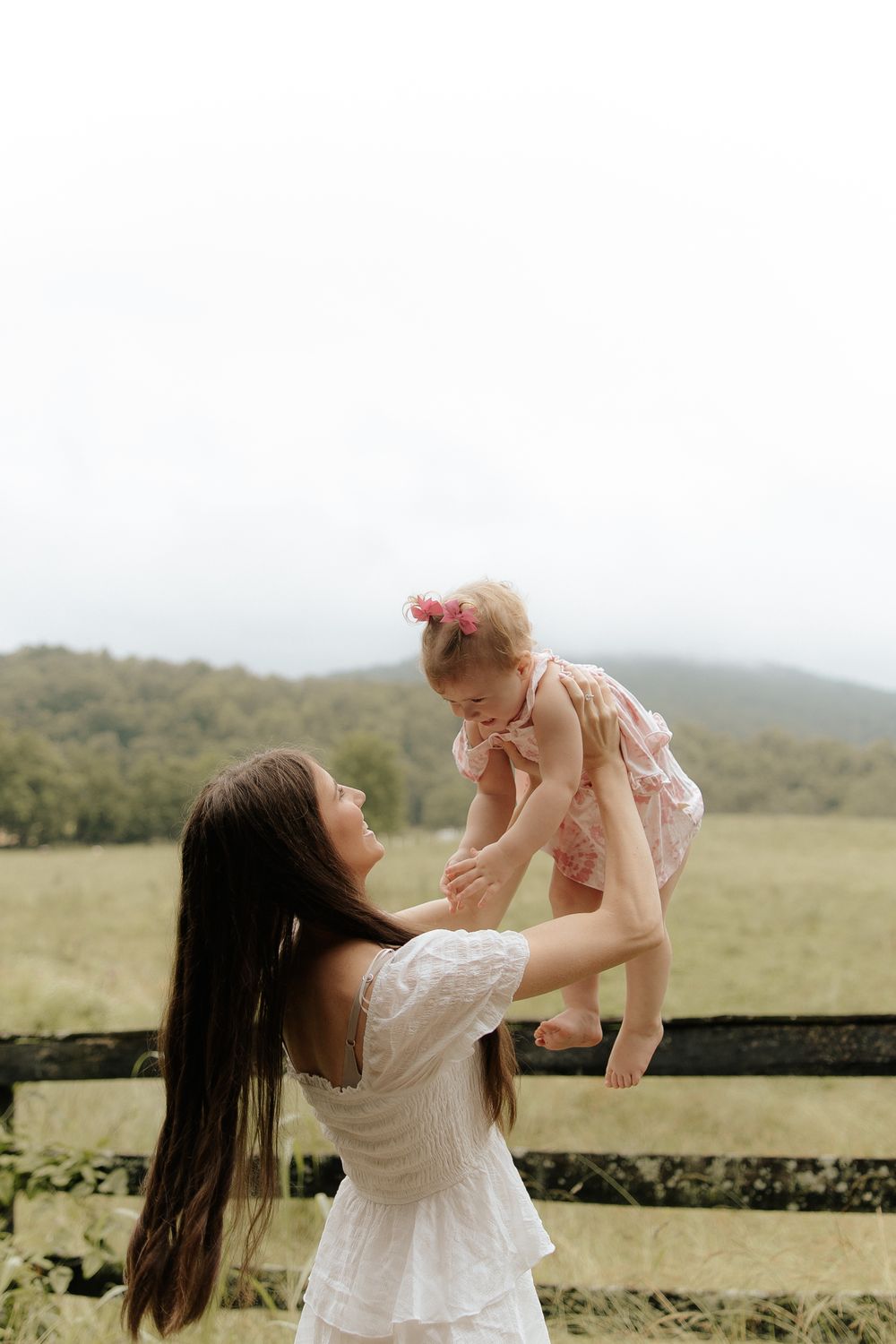 A loving moment captured between parent and child playing outdoors on a rustic fence against rolling hills.