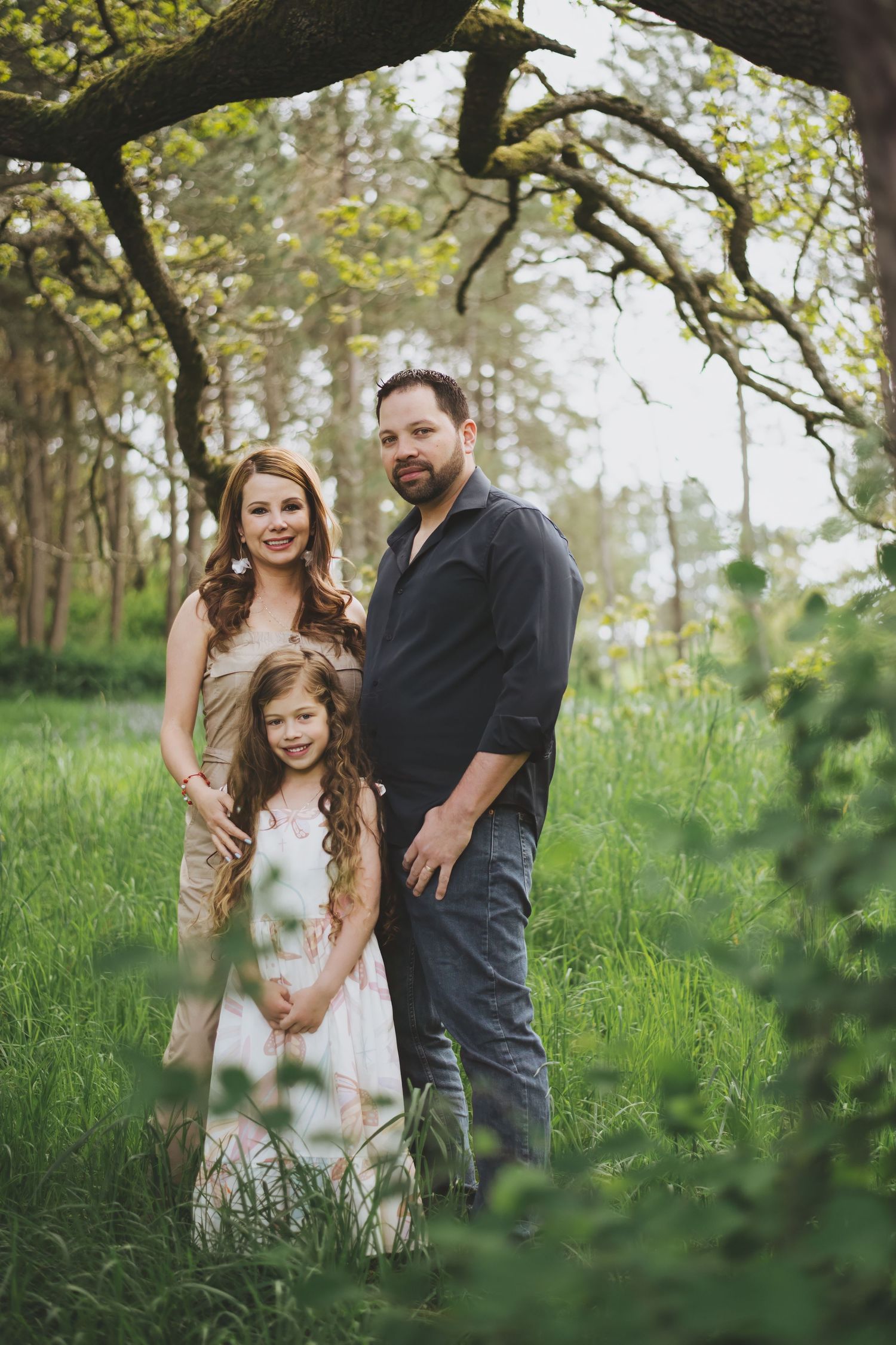 A family poses together in a lush green spring forest setting under tree branches with soft natural lighting.