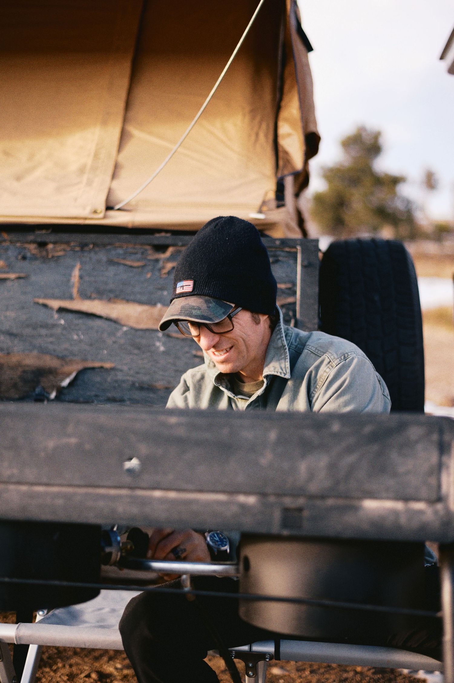 Person in black beanie hat and glasses working at outdoor setup with camping gear at golden hour.