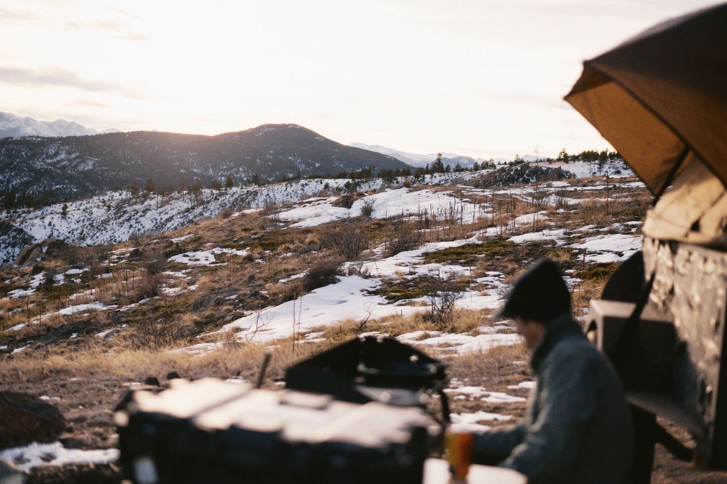 A snowy mountain landscape at sunset with a figure silhouetted against the golden sky beneath an umbrella.