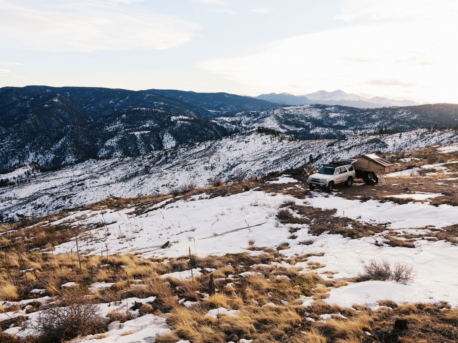 Snow covered mountain landscape with patches of dry grass and rock formations against a cloudy winter sky.
