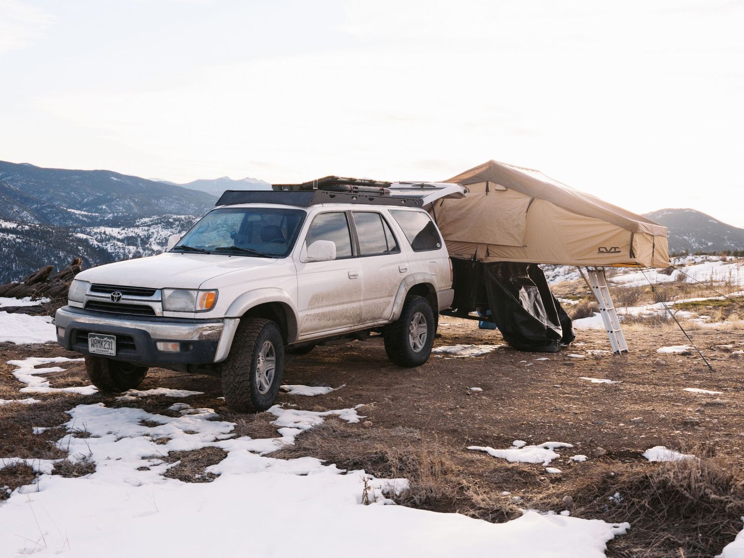A white Toyota 4Runner parked next to a tan rooftop tent setup in a snowy mountain camping area.