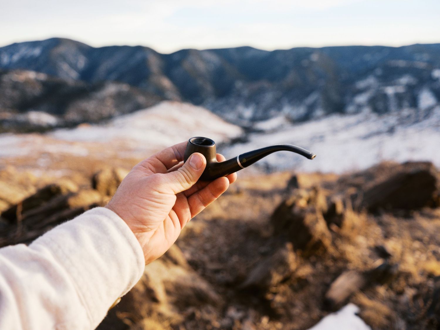 A hand holds tobacco pipe against a backdrop of rugged mountain terrain on a clear day.