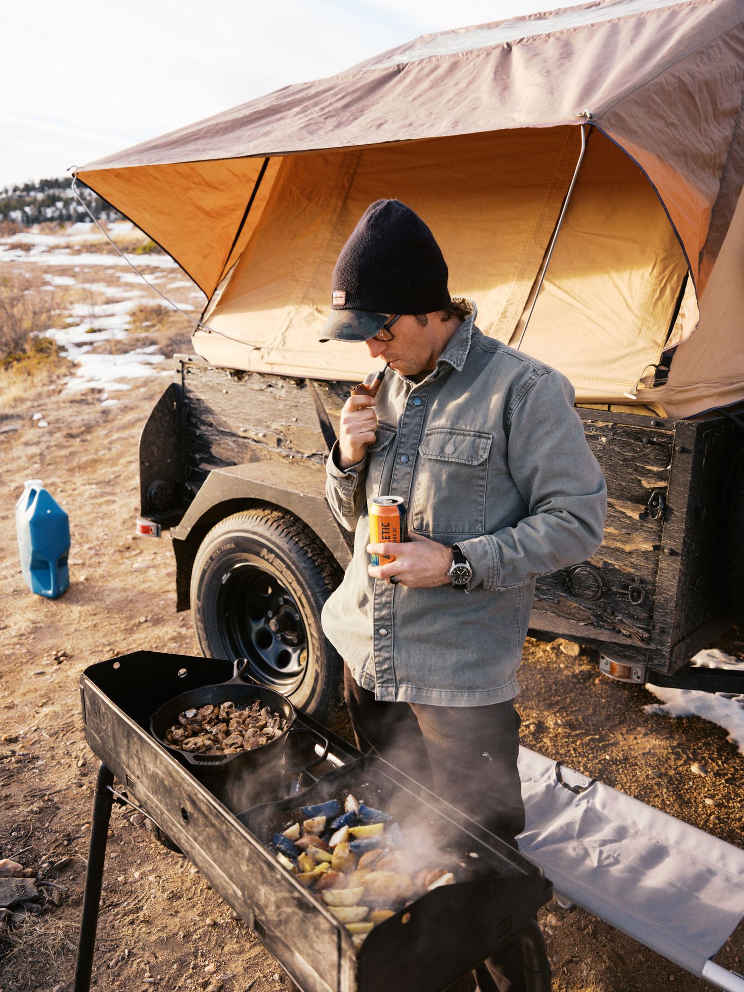 A camper cooks breakfast on a grill beneath a rooftop tent canopy during a winter camping trip in snowy terrain.