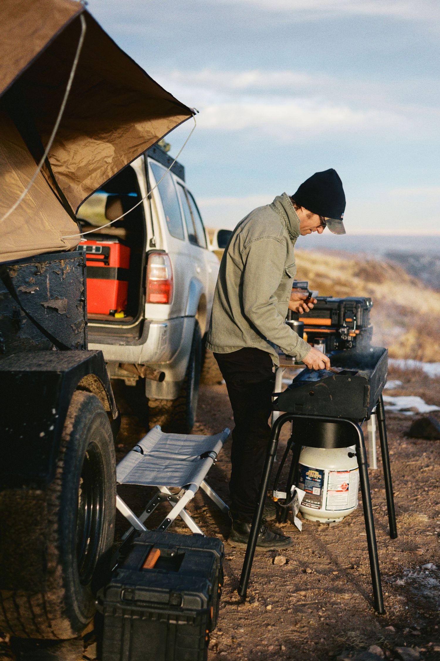 Camping enthusiast cooking on portable stove next to SUV with rooftop tent in remote mountainous location.