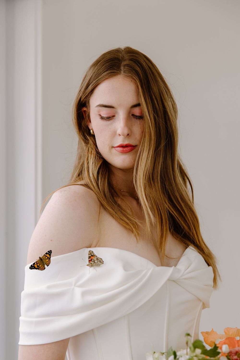 Monarch butterfly perches on elegant white off-shoulder dress as woman with long blonde hair poses against light wall.
