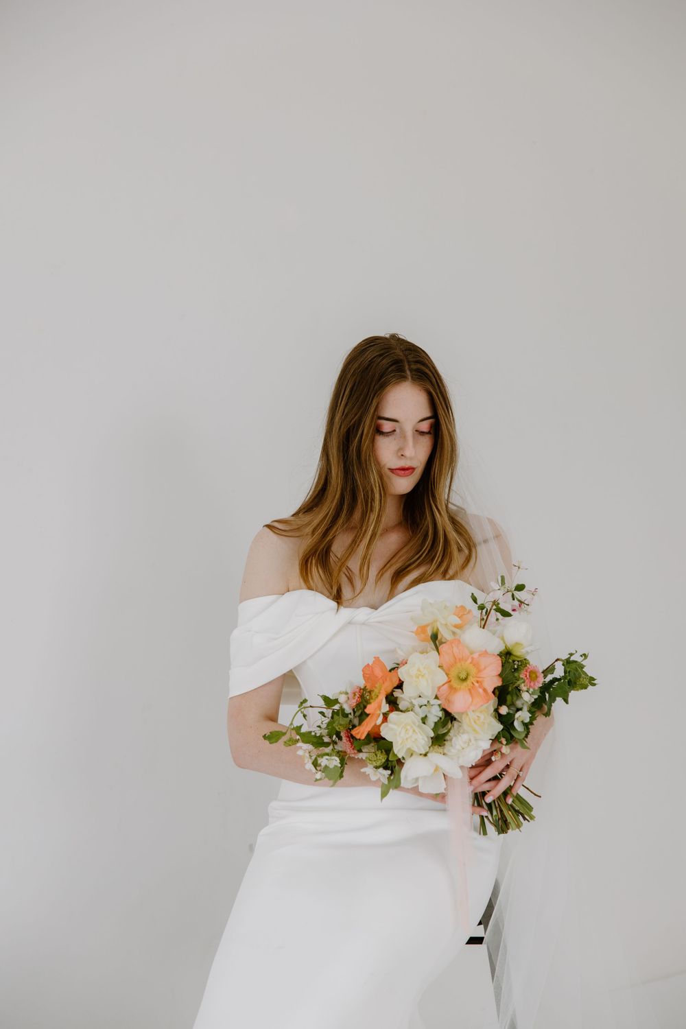 Off-shoulder white wedding gown and delicate peach and cream floral bouquet against minimalist light background.